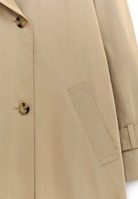 Trench-coat beige avec un grand bouton, une poche latérale et des coutures nettes. Le tissu présente une texture lisse et une coupe ajustée.