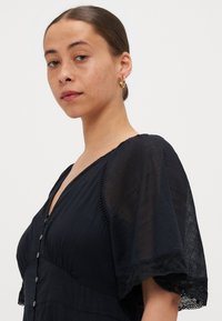 Robe noire avec des manches courtes et légères, en tissu texturé, décolleté en V, et détails de boutons sur le devant. Finition en dentelle délicate aux bords des manches.