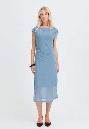Femme blonde portant une robe midi bleu clair à manches courtes et ourlet transparent, chaussures pointues marron foncé et un bracelet noir, debout sur un fond blanc.