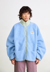 Lichtblauwe fleece jas met groene drukknoopkraag, twee voorzakken en een gestructureerde stof. Draagt over roze broek.