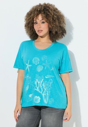 Kobieta z kręconymi włosami, ubrana w turkusowy t-shirt z białymi ilustracjami morskiego życia, w tym rozgwiazd, muszli i korali, oraz ciemne dżinsy.