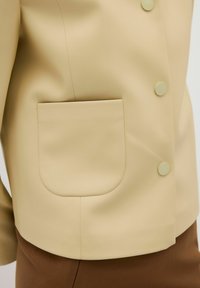 Helles beiges Jacket mit glatter Textur, ausgestattet mit zwei runden Knöpfen und einer abgerundeten Tasche an der Seite. Brauner Stoff ist darunter sichtbar.