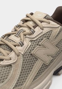 Κοντινό πλάνο μπεζ και καφέ αθλητικού παπουτσιού New Balance με διχτυωτό ύφασμα, σουέτ επενδύσεις και μπεζ κορδόνια με ριγέ θηλιές κορδονιών.