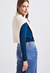 Femme aux longs cheveux portant un haut à manches longues bleu, un jean bleu et un gilet sans manches fuzzy crème, devant un fond uni.