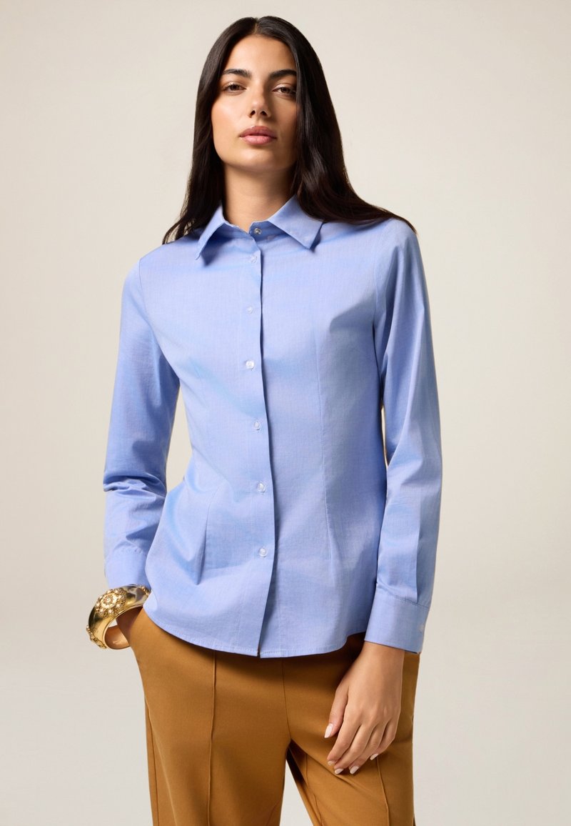 Camicia azzurro chiaro con colletto a punta, maniche lunghe e vestibilità su misura, abbinata a pantaloni marroni. Tessuto liscio e design semplice.