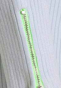 Ljust blå ribbad tröja med en silverdragkedja och en neongrön detalj längs knäppningen. Texturerad tyg med fina detaljer.