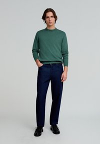 Grüner, gestrickter Pullover mit Rundhalsausschnitt, kombiniert mit dunkelblauen, geraden Jeans. Schwarze Schnürschuhe runden das Outfit ab.