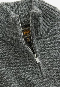 Pull en tricot gris avec un col haut et une fermeture éclair quart de zip. Présente un motif texturé et un zip métallique avec une étiquette à l'intérieur.