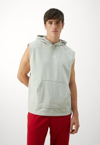 Grå ærmeløs hættetrøje med en frontlomme og snørehætte. Har et lille hvidt logo på brystet. Stoffet virker blødt og afslappet.