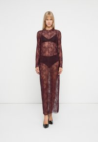 Sieviete, kas valkā garu, caurspīdīgu bordo mežģīņu kleitu virs atbilstošā krūštura un apakšbiksēm, stāv uz vienkārša balta fona, melnās kurpes ar augstu papēdi.