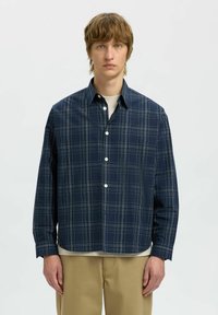 Camicia a quadri blu navy con maniche lunghe, colletto button-down e bottoni bianchi, sovrapposta a un capo base di colore chiaro.