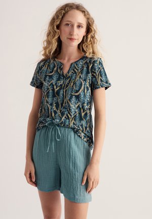 Kvinde med krøllet blondt hår iført en blå mønstret kortærmet top og teksturerede lyseblå shorts med snor, står mod en ensfarvet baggrund.