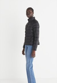 Črna puhovka z vodoravnimi šivi, visokim ovratnikom in tesnim prilegajanjem. Nosi se čez modre kavbojke, ki imajo gladko, sijočo teksturo.