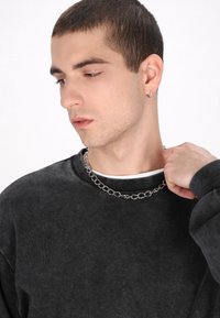 Schwarzer Sweatshirt mit gerippten Bündchen und Halsausschnitt, über einem weißen Hemd getragen, kombiniert mit einer silbernen Kettenhalskette. Glatte Textur.