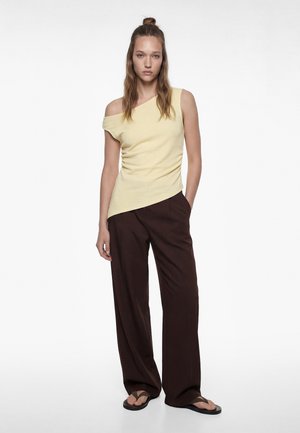 Frau trägt ein hellgelbes Ein-Schulter-Oberteil, dunkelbraune weit geschnittene Hosen und schwarze Flip-Flops, steht mit einer Hand in der Tasche.