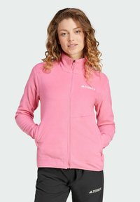 Chaqueta de forro polar rosa con un cuello alto, cremallera completa y bolsillos laterales. Presenta un logo de Terrex en el pecho izquierdo. Textura suave, corte relajado.
