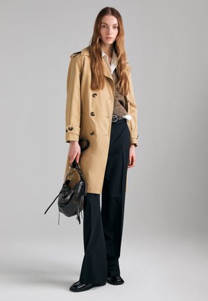 Donna con trench beige, maglione marrone, camicia bianca, pantaloni neri a gamba larga, che tiene una borsa nera in pelle, in piedi su sfondo grigio.