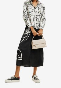 Hvid skjorte med knapper og farverige abstrakte ansigtsdesigns, sort nederdel med hvide kanter, beige håndtaske og sorte sneakers med hvide såler.