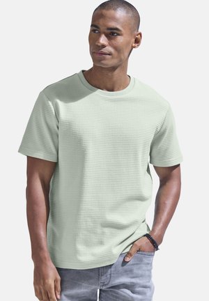 Helles grünes T-Shirt mit kurzen Ärmeln und einem strukturierten, waffelartigen Muster; Rundhalsausschnitt-Design; kombiniert mit hellgrauen Jeans.