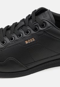 Black leather sneaker with a smooth finish, featuring round laces, metal eyelets, a subtle logo in rose gold, and perforated accents.