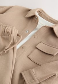 Veste décontractée beige et pantalon à cordon assorti superposés sur une chemise blanche côtelée, présentés à plat avec boutons visibles et détails de poche.