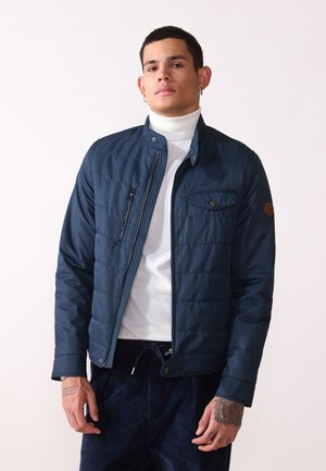 Chaqueta acolchada en color navy con cierre de cremallera, cuello alzado y bolsillo en el pecho. Presenta una textura suave y un sutil logo marrón en la manga.