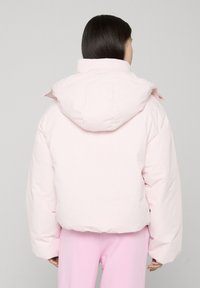 Persoon met lang donker haar draagt een lichtroze pufferjack met capuchon en bijpassende roze broek, staand tegen een effen grijze achtergrond.