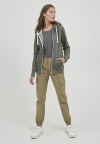 Hoddie zippé vert chiné avec des accents blancs, associé à un T-shirt gris et un pantalon jogger beige doté de poches cargo sur les côtés et de poignets élastiques.