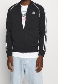 Black zip-up jacket made of fabric, featuring white stripes on sleeves and an embroidered logo on the chest. Worn over a white shirt.