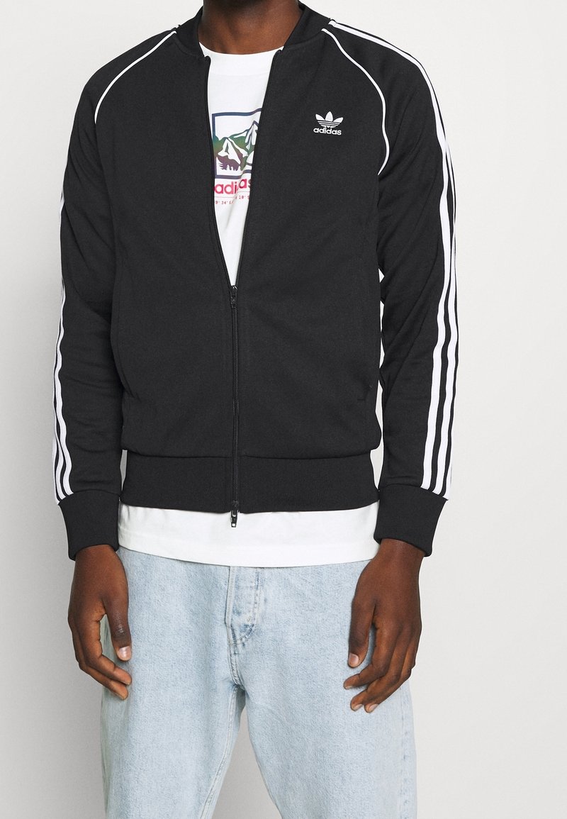 Black zip-up jacket made of fabric, featuring white stripes on sleeves and an embroidered logo on the chest. Worn over a white shirt.