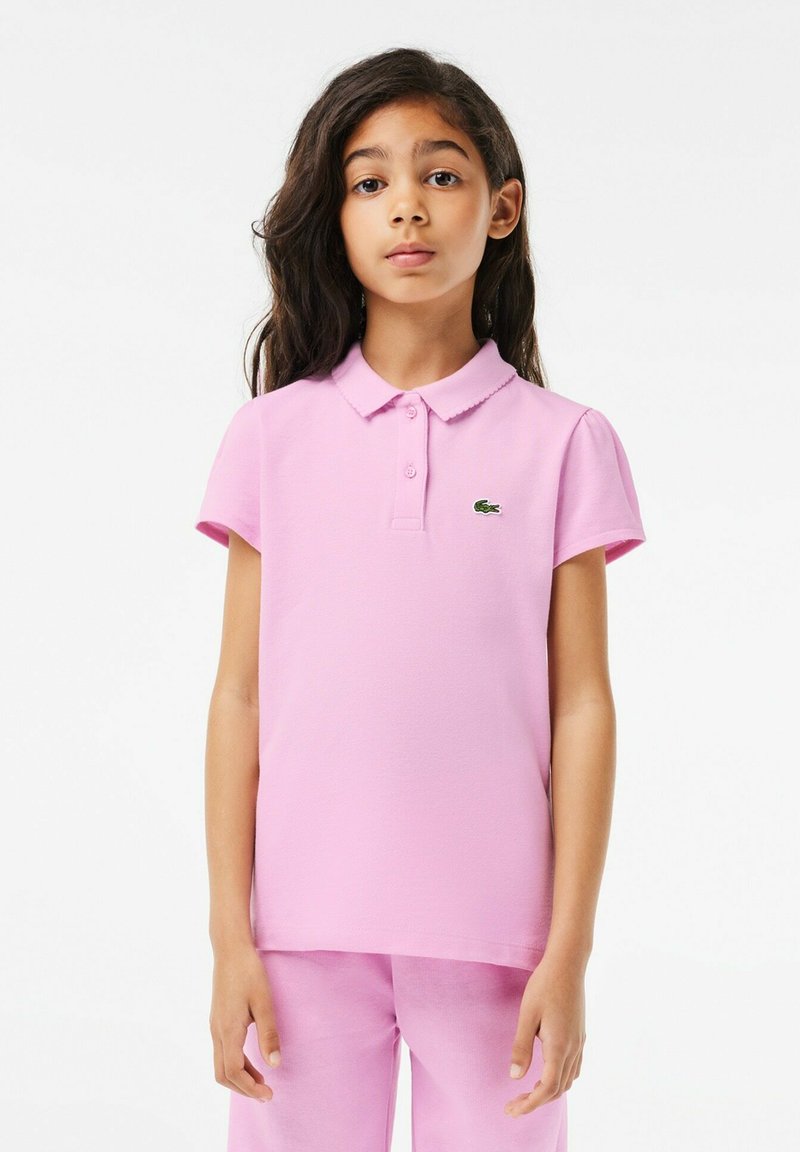 Polo rose avec col et manches courtes, fabriqué en tissu léger, arborant un petit logo de crocodile vert sur la poitrine.