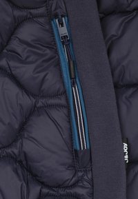 Detail van een marineblauw gewatteerd jasje met een blauwe rits en zwarte hardware, en een gebreid stoffen accent op de kraag. Textuur van het materiaal is zichtbaar.