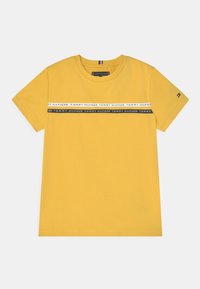 Żółta bawełniana koszulka z krótkim rękawem, z poziomym pasem z napisem "TOMMY HILFIGER" w czerni i bieli na klatce piersiowej.