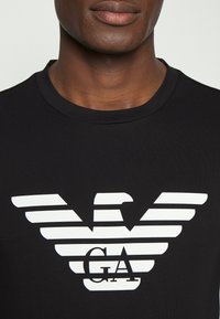 Svart bomull t-shirt med en framträdande vit logotypdesign med horisontella linjer och texten "GA", rund halsringning och korta ärmar.