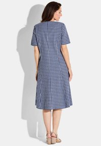 Abito a scacchi blu navy e bianco, maniche corte, silhouette svasata, lunghezza ginocchio, vista posteriore con leggere pieghe e abbinato a tacchi beige.