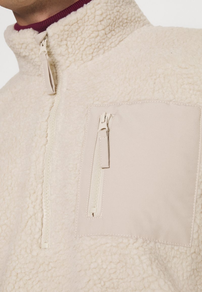 Cremefarbener Fleece-Pullover mit hohem Kragen, ausgestattet mit einer Vordertasche mit Reißverschluss. Glatte Textur mit einem weichen, flauschigen Design.