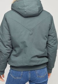 Graue isolierte Jacke mit Kapuze, aus strukturiertem Stoff und schrägem Nahtdesign auf der Rückseite; kombiniert mit blauen Jeans.