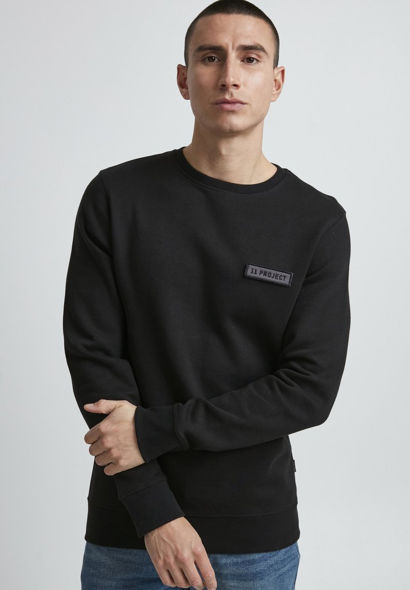 Black crewneck sweatshirt made of cotton blend, featuring a textured patch with "11 PROJECT" logo on the chest and ribbed cuffs and hem.