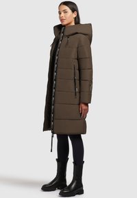 Manteau d'hiver marron avec capuche, présentant des panneaux matelassés verticaux, une fermeture éclair intégrale et des poches latérales. Porté sur des leggings noirs et des bottes.