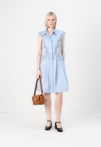 Vestito camicia azzurro chiaro con dettagli ricamati a onde, maniche corte e chiusura con bottoni. Accessoriato con una borsa a tracolla marrone.