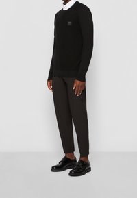 Svart stickad tröja med en liten applikation på bröstet, tillsammans med skräddarsydda svarta byxor och blanka svarta skor. Enkelt, modernt design.