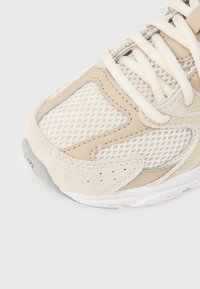 Tuvplāns ar bēša un balta sporta apavu purngalu, tīkla audumu, zamšādas uzlikām un auklām uz gaiša fona.