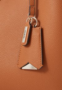 Tan textured handbag featuring a prominent silver logo tag and a triangular metal accent. The leather has a pebbled finish.