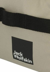 Beige canvas tas met een gestructureerd oppervlak. Voorzien van een horizontale zwarteriem en een rechthoekig zwart label met witte tekst: "Jack Wolfskin."