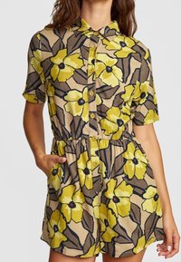 Combinaison à manches courtes en beige avec un grand motif floral jaune et des accents noirs ; dotée d'une fermeture à boutons à l'avant et d'une taille élastique avec poches.