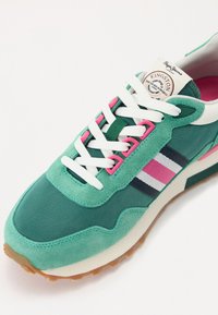 Grønn idrettssko med en blanding av semsket skinn og stoff, hvite lisser, rosa detaljer og et stripet design i marineblått, rosa og hvitt.