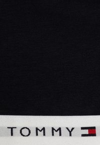 Tissu noir avec une taille blanche portant l'inscription "TOMMY" en noir, accompagnée d'un détail logo rectangulaire rouge et bleu. Texture lisse.