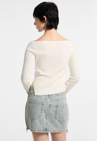 Top a maniche lunghe a coste color crema con scollo ampio, abbinato a una mini gonna in denim blu chiaro con orli sfilacciati e chiusure con zip.