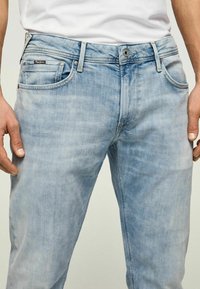 Jeans de denim azul claro con un acabado desgastado, que presentan un diseño clásico de cinco bolsillos, cierre de botón y una sutil etiqueta con el logotipo en la parte frontal.