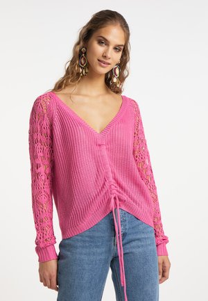 Frau trägt einen rosa Strickpullover mit Spitzeärmeln und einem Kordelzug vorne, kombiniert mit blauen Jeans, steht vor einem schlichten Hintergrund.